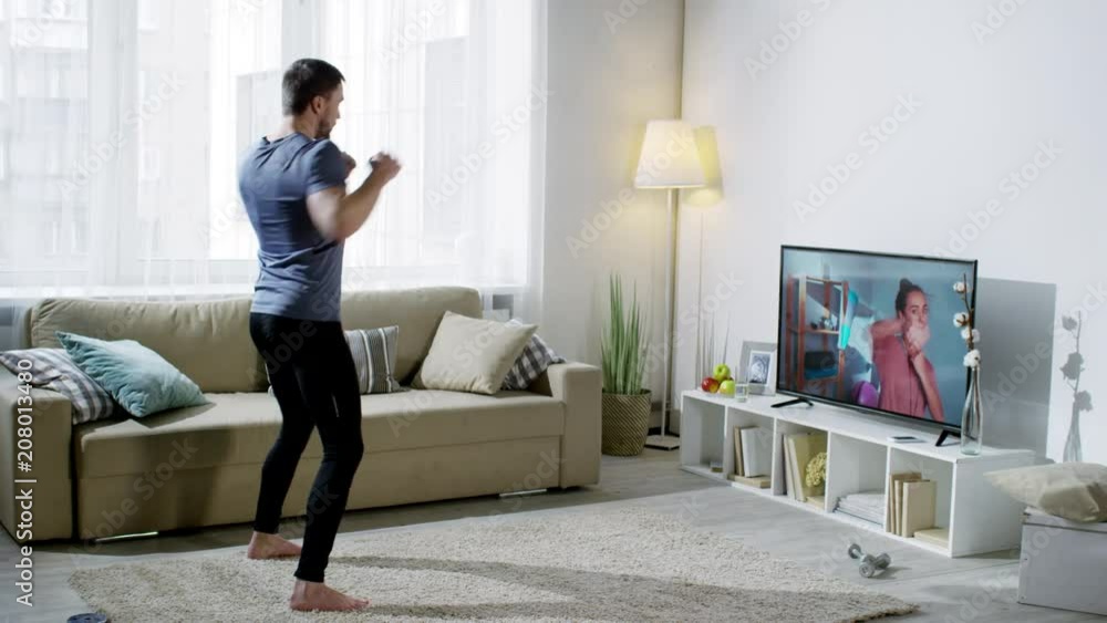 Wide shot of man in sportswear looking at online personal trainer on TV screen and doing boxer squat punch exercise at home Wide shot of man in sportswear looking at online personal trainer on TV screen and doing boxer squat punch exercise at home