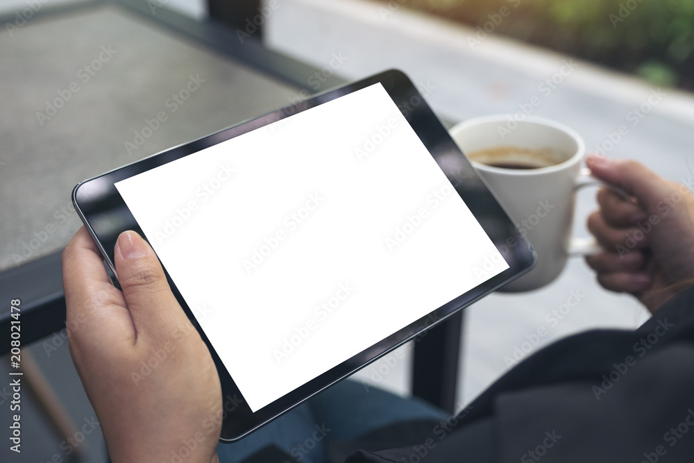 Mockup image of a woman sitting cross legged and holding black tablet pc with blank white desktop screen while drinking coffee in cafe Mockup image of a woman sitting cross legged and holding black tablet pc with blank white desktop screen while drinking coffee in cafe