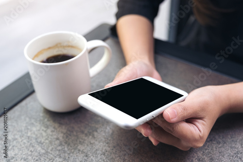 Mockup image of hands holding white mobile phone with blank black desktop screen with coffee cup on table in cafe