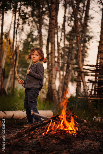 child girl standing at campfire with stick. Burning fire in the woods at night or evening