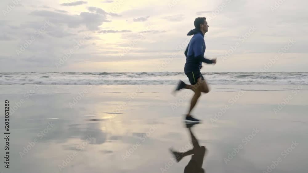 Side view of sporty man in hoodie and shorts running on sand and listening to music when warming up before summer workout at the beach Side view of sporty man in hoodie and shorts running on sand and listening to music when warming up before summer workout at the beach