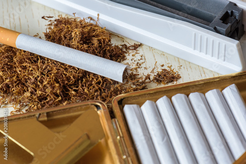 A pile of natural tobacco on a white wooden table, a device for manual production of cigarettes and cigarettes in a cigarette case. Smoking. Nicotine addiction. Health hazard.