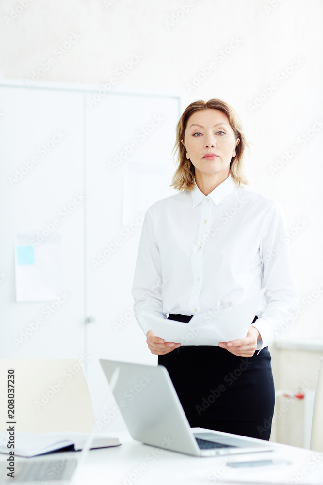 Businesswoman in formal wear standing in office room holding papers near table with laptops and looking at camera Businesswoman in formal wear standing in office room holding papers near table with laptops and looking at camera