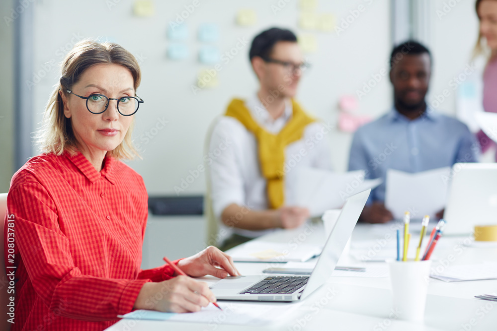 Serious businesswoman using laptop and writing at table in office with people on background Serious businesswoman using laptop and writing at table in office with people on background