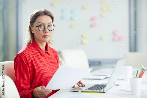 Canvas Print Adult emotionless woman in glasses sitting with paper at table using laptop and looking at camera