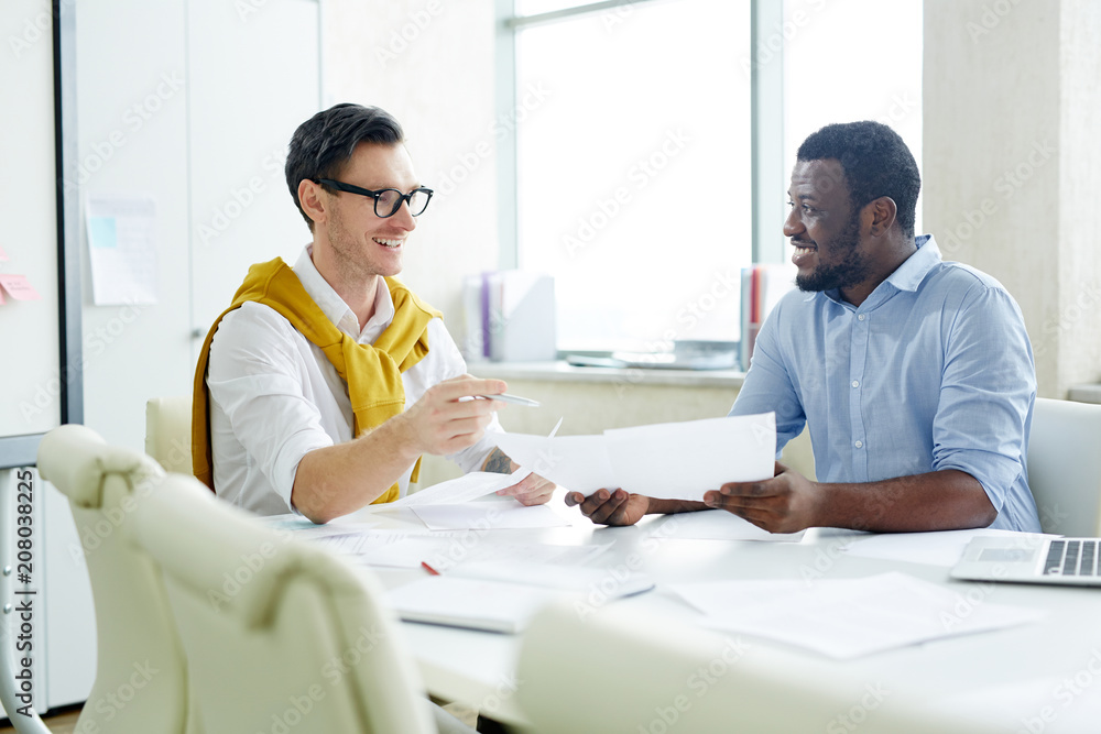 Cheerful stylish multiracial men chatting at table in light office and discussing papers while working. Cheerful stylish multiracial men chatting at table in light office and discussing papers while working.