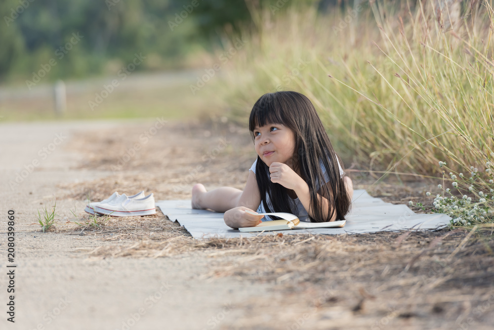 A little girl is lying on her stomach reading on the grass. She has a look of enjoyment on her face and she looks very relaxed. There is quite a bit of negative space around her.