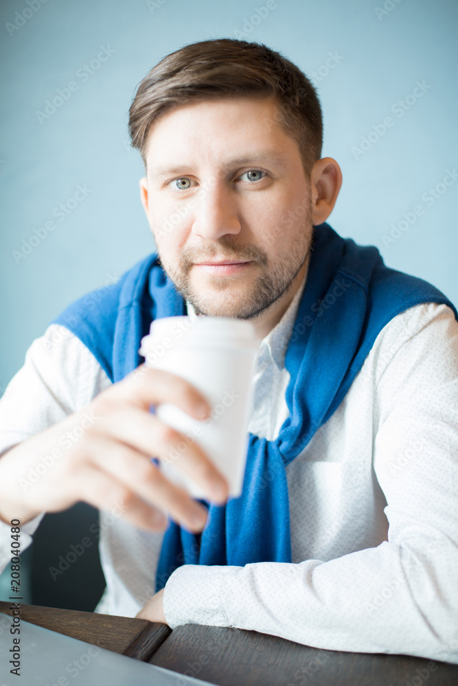 Handsome man in elegant outfit holding cup of hot drink and looking at camera while sitting near light blue wall. Handsome man in elegant outfit holding cup of hot drink and looking at camera while sitting near light blue wall.