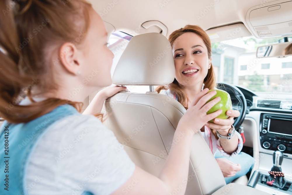 side view of daughter giving fresh apple to smiling mother in car side view of daughter giving fresh apple to smiling mother in car