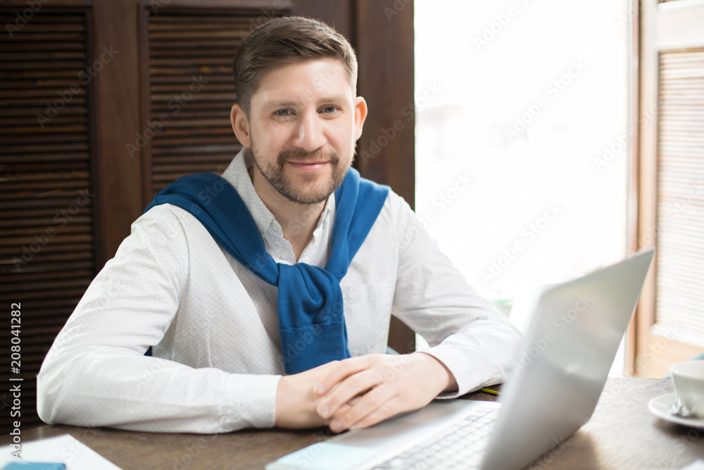 Handsome man in elegant clothes smiling and looking at camera while sitting at cafe table near laptop. Handsome man in elegant clothes smiling and looking at camera while sitting at cafe table near laptop.
