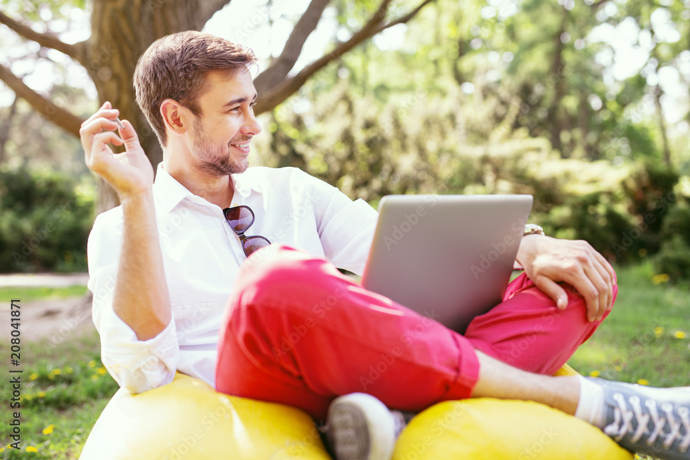 Fototapeta premium Making money online. Cheerful stylish man sitting in the open air and holding a coin