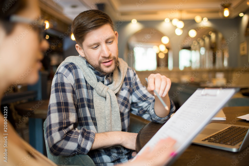 Handsome man in casual clothes sitting at cafe table and pointing at important items in clipboard document for colleague. 