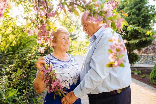 Beautiful senior couple in love looks at each other under pink blooming tree in sunset outside in spring garden nature.