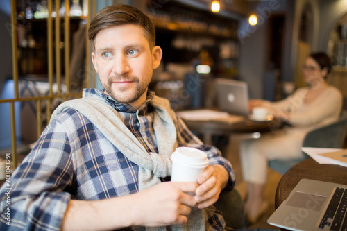 Handsome man in casual clothes holding cup of tasty hot beverage and looking away while sitting at cafe table near laptop. 