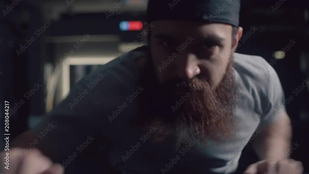 Brutal and powerful man with a beard, confidently looking forward, intensively moving his hands and doing an exercise on the ellipsoid simulator. Heavy physical activity. Portrait in dark background. Brutal and powerful man with a beard, confidently looking forward, intensively moving his hands and doing an exercise on the ellipsoid simulator. Heavy physical activity. Portrait in dark background.