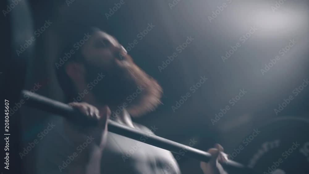 A brutal man with a beard is engaged in weightlifting in a dark room and performs the taking of the bar on his chest. Crossfit, weightlifting. The man is focused on doing sports and training. A brutal man with a beard is engaged in weightlifting in a dark room and performs the taking of the bar on his chest. Crossfit, weightlifting. The man is focused on doing sports and training.