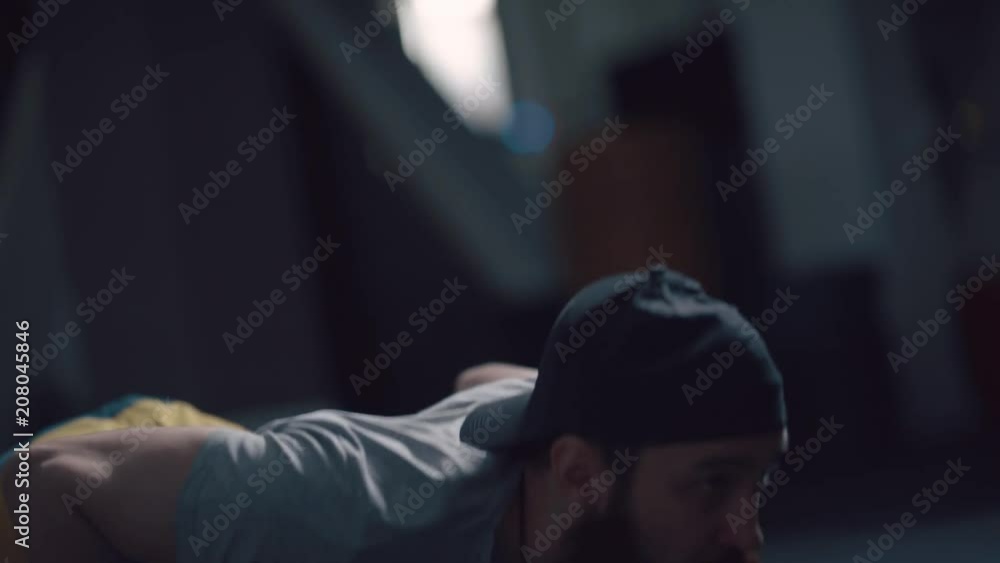 Strong man with a beard and in a cap wringing out (push-up), lying on the floor. He looks in front of him, technically performs the exercise. Dark background, profile of the athlete. Strong man with a beard and in a cap wringing out (push-up), lying on the floor. He looks in front of him, technically performs the exercise. Dark background, profile of the athlete.