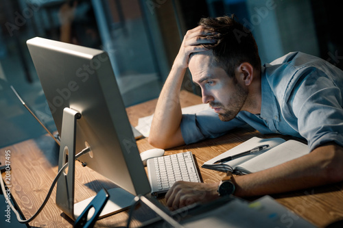 Obraz na plátně Young man leaning on hand looking extremely tired while finishing overtime project watching computer at table