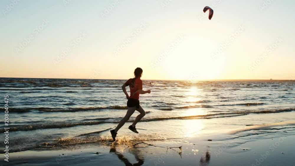 Man runs along the beach and is happy at the finish on the background of kitesurfers at sunset. Athlete splashes water Man runs along the beach and is happy at the finish on the background of kitesurfers at sunset. Athlete splashes water