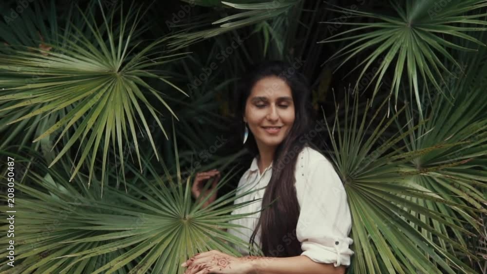 Young woman with dark hair is standing in tropical garden. She is looking on huge beautiful leaves of palm and stroking it Young woman with dark hair is standing in tropical garden. She is looking on huge beautiful leaves of palm and stroking it