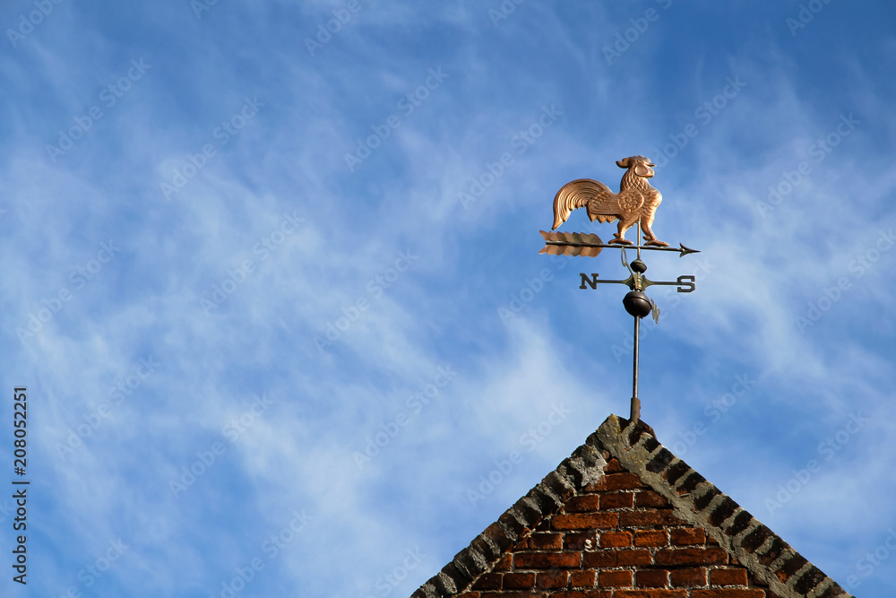 Weathercock (also known as weather vane or wind vane) on top of old red brick construction against a blue sky with clouds during a sunny day. Weathercock (also known as weather vane or wind vane) on top of old red brick construction against a blue sky with clouds during a sunny day.
