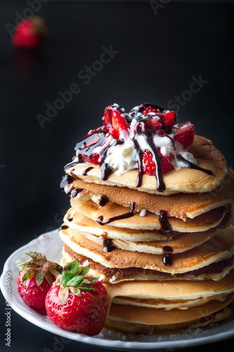 Stack of pancakes with strawberries, whip cream and chocolate syrup on a white plate on a black background. Copy space.