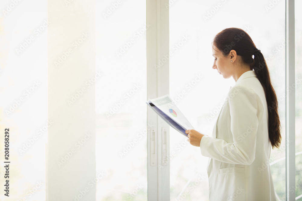 Business woman standing working near a window and holding clipboard at office. Business woman standing working near a window and holding clipboard at office.