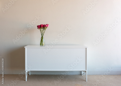 Variegated red roses in glass vse on white sidetable against neutral wall background photo