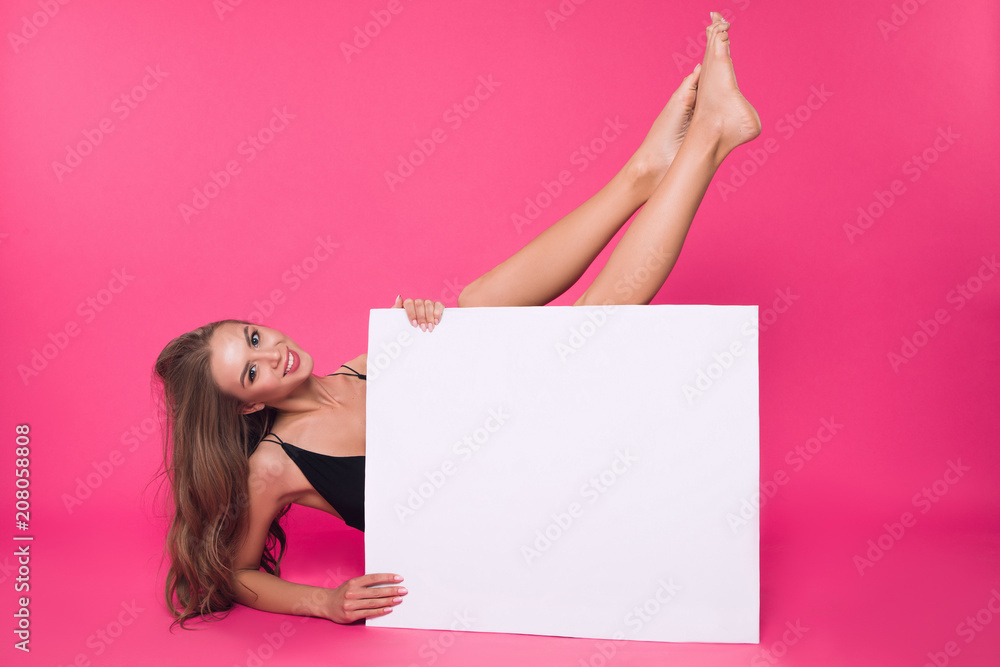 Naklejka premium In a mood of total nudity! Attractive cheerful well-graced lady pleasantly smiling, while holding up her legs, while sitting on the floor with white board.