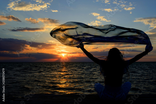 A young slender girl stands on a wooden bridge (fishing bridge) spreading her hands over the sea and looking at the sunset (dawn) of the sun. Life and freedom. 