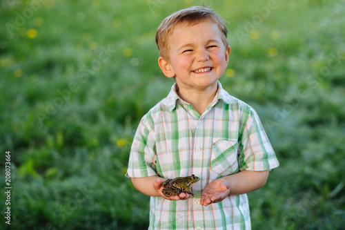 Cute blond little boy with defferent emotions holding little frog at summer day over green background. photo