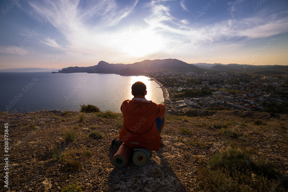 Naklejka premium man sits on top of a hill and looks at the sun set near the sea