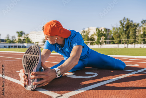 Young athlete male stretching his leg on a track in stadium, preparing for running and jogging workout. Caucasian man exercising outdoors wearing blue sportswear. Sport, people, lifestyle
