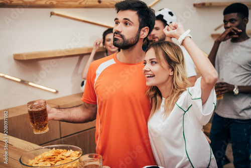 young couple with multicultural friends celebrating and watching football match at bar