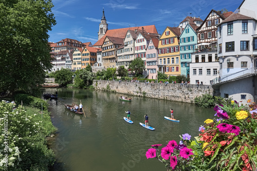 Picturesque view of historical center of Tubingen, Germany. Unidentified people ride along the Neckar river on the traditional punt boats and standup paddle boards, and have a rest at the embankment.