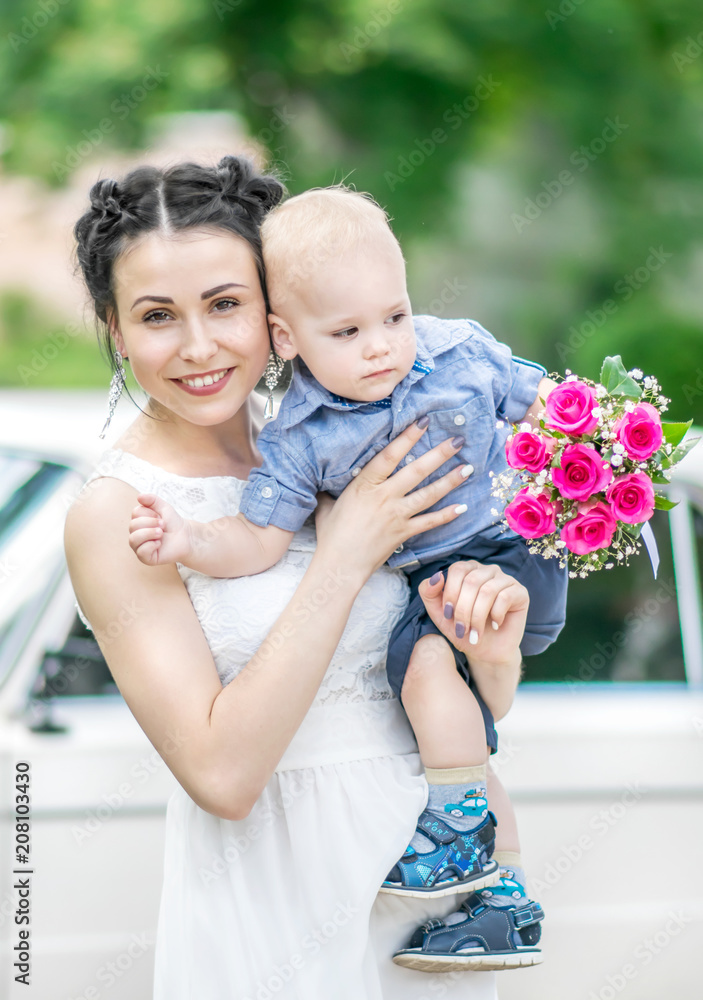 Portrait of pretty young female bride walking, holding baby boy with wedding roses bouquet and looking into camera at sunny summer park. Mother and her little son on her wedding. Portrait of pretty young female bride walking, holding baby boy with wedding roses bouquet and looking into camera at sunny summer park. Mother and her little son on her wedding.