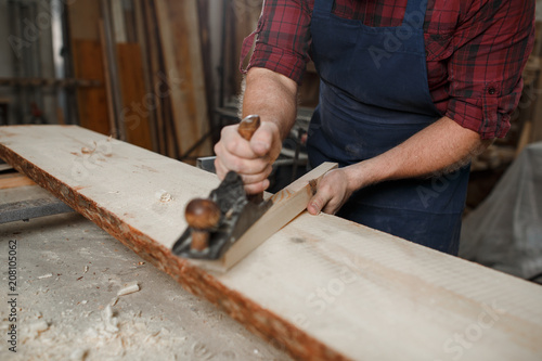 Master carpenter in shirt and apron strokes plane in workshop. Close up