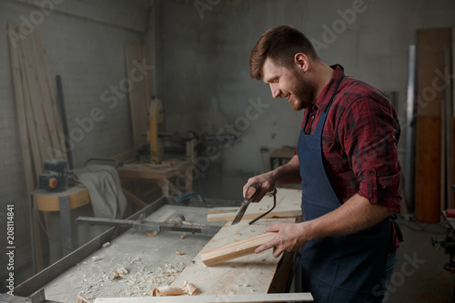 Master carpenter in shirt and apron works as an ax in workshop