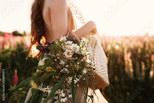 Woman holding wildflowers bouquet in straw bag, walking in flower field on sunset. Horizontal noface still life