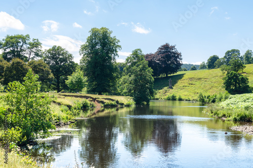 View of a river in summertime as sheep graze in the middle ground. Photo of the Derwent River at Chatsworth Park in the Peak District, Derbyshire, England  photo