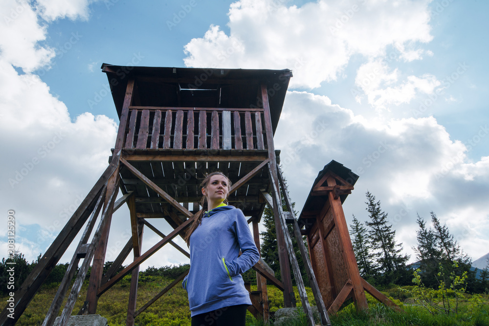 Beautiful young woman in sportswear standing near the wooden observation structure in mountains on sunny spring day. Hiking. Carpathian Mountains, Ukraine