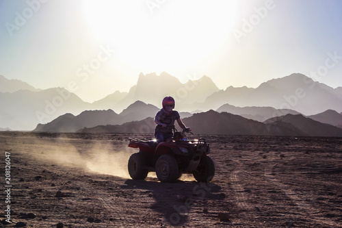 Young man rides an ATV on the desert over background of mountains at sunset