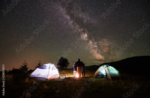 Back view of mother and two sons backpackers resting at camping in mountains, standing beside campfire and two illuminated tents, looking at night sky full of stars, Milky way, enjoying night scene.