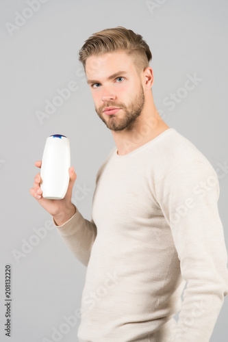 Man confident face holds shampoo bottle, grey background. Guy with bristle holds bottle shampoo, copy space. Hair care and beauty supplies concept. Man enjoy freshness after washing hair with shampoo