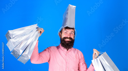 Man with trendy beard and smiling face isolated on blue background. Hipster in pink shirt doing shopping, big spender. Happy bearded man carrying silver bags and wearing one as hat, shopping concept