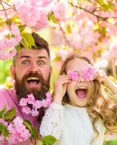 Father and daughter on happy face play with flowers as glasses, sakura background. Girl with dad near sakura flowers on spring day. Child and man with tender pink flowers in beard. Family time concept