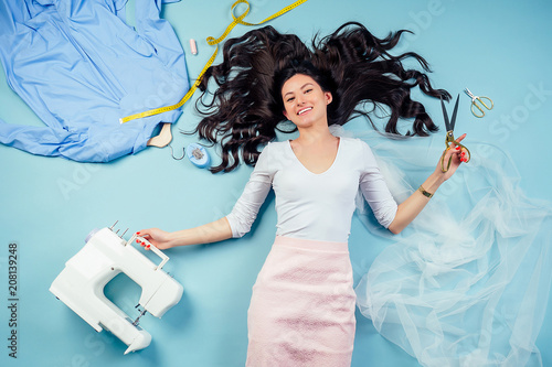 brunette woman seamstress tailor dressmaker lying (fly) on the floor with sewing machine, coats on a hanger and measuring tape on a blue background in studio view from top. inspiration and creativity photo