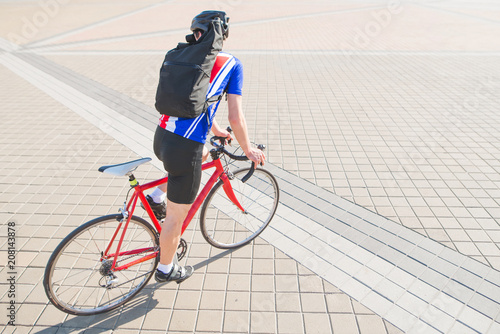 Cyclist with a backpack on his back rides a red bike on a square with a pavement. A man in sportswear and a helmet rides a bicycle in the city