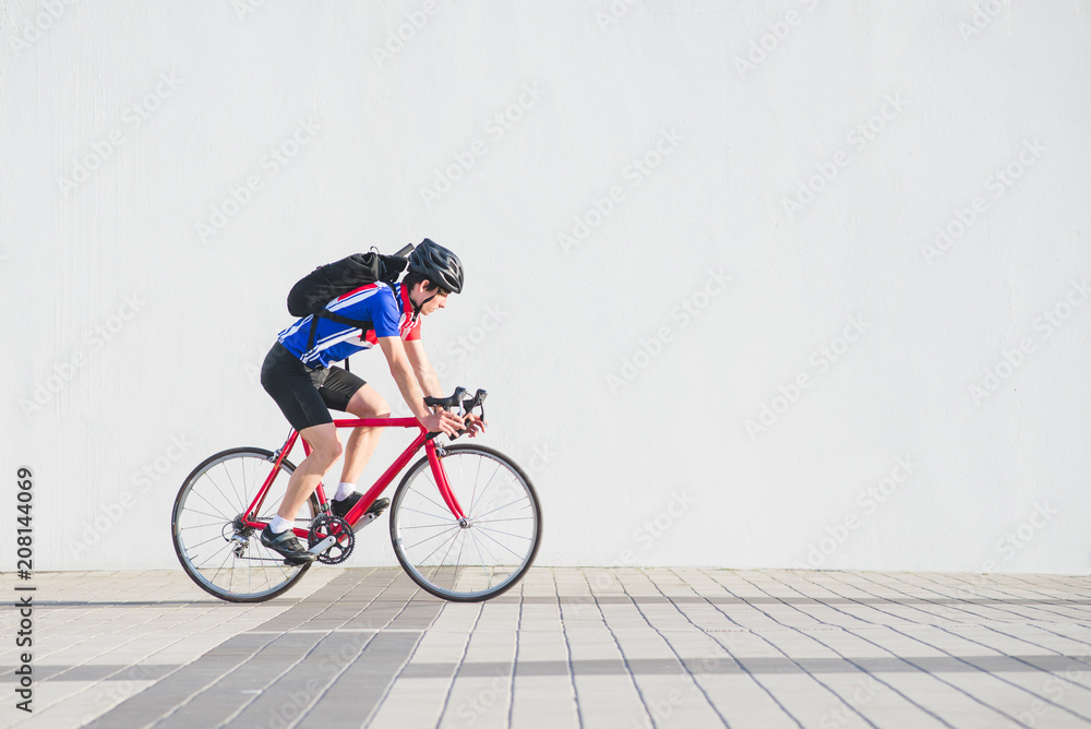Obraz premium Cyclist in a helmet, athletic cycling clothes and a backpack rides on the background of a white wall. Athlete bicyclist side view on the background of a light wall. Sports concept. Copyspace