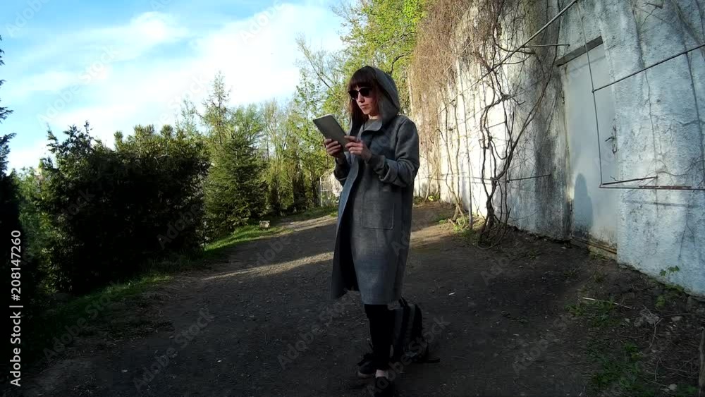 pretty brunette in a gray cardigan using digital tablet at the park in front of the concrete wall with dry vine pretty brunette in a gray cardigan using digital tablet at the park in front of the concrete wall with dry vine